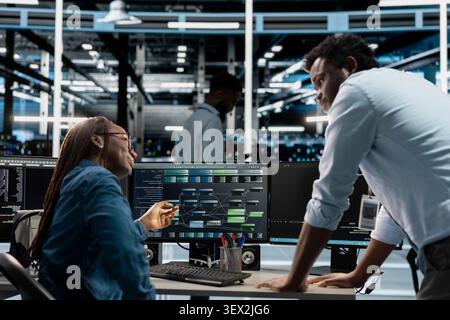 African American supervisor checks an engineer programming in a server hub, overseeing repairs on data center racks and infrastructure. Stock Photo