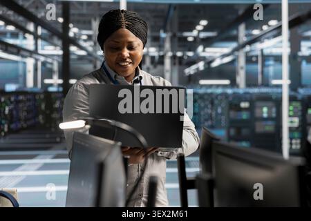 Smiling data center engineer uses a laptop to review server infrastructure, diagnostics and AI technology in a data center. Stock Photo