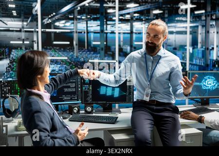 Coworkers coding in high tech server room sharing a fist bump, pleased after updating AI automation scripts, while happy server room team members celebrating programming AI algorithms. Stock Photo