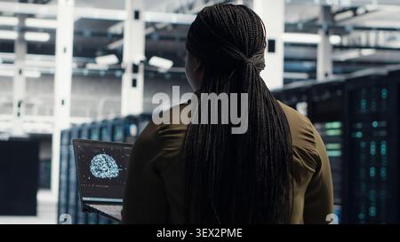 Programmer uses laptop-based deep learning tools in a server room to improve AI operations and system efficiency. Stock Photo