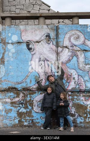 Octopus mural dominates weathered concrete wall. Two children and an adult pose nearby, smiling. Their playful stance adds charm to the urban scene. B Stock Photo