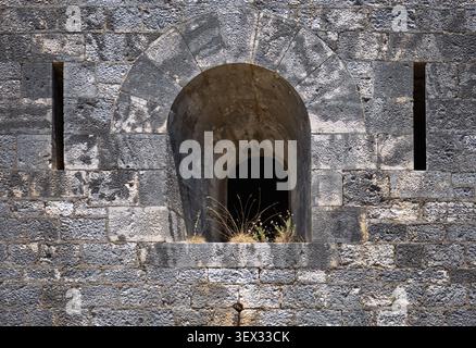 ancient stone fortress wall with an arched embrasure window and vertical arrow slits, featuring weathered grey bricks and dry grass growing on the led Stock Photo
