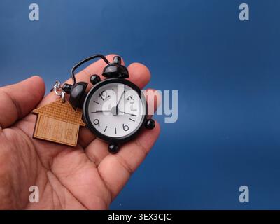 A hand holds a small black vintage alarm clock next to a wooden house-shaped keychain on a blue background. Stock Photo
