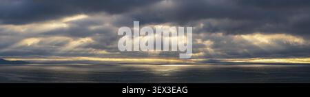 Dramatic sunbeams breaking through storm clouds over ocean, showing a moody coastal seascape with atmospheric light. Stock Photo