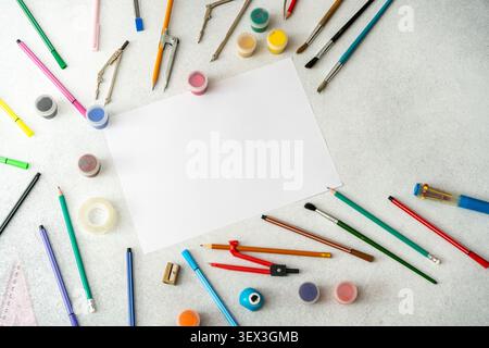 Art Workspace desk featuring a blank white sheet of paper, colorful paints pencils, brushes, and drafting tools. School study background top view table Stock Photo