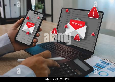 Hand holds smartphone beside laptop showing red warning email alerts, phishing message icons and blurred inbox screens for cybersecurity threat detect Stock Photo