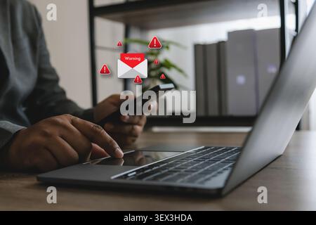 Hand holds smartphone beside laptop with warning email alert icons, phishing message notice, spam notification and cybersecurity risk concept for onli Stock Photo