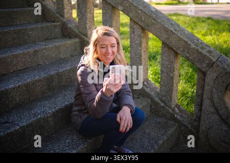Woman sitting on old stone stairs in a park, smiling while looking at her smartphone Stock Photo