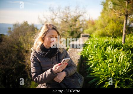 Blonde woman scrolling through her smart phone in a sunny park with trees Stock Photo