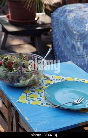 Fresh mixed salad on rustic outdoor table with empty blue plate in a cozy garden dining setting Stock Photo
