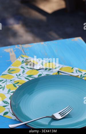 Empty blue ceramic plate with fork on rustic outdoor table setting and lemon patterned napkin Stock Photo