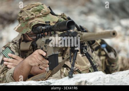 Military sniper aiming precision rifle during tactical reconnaissance mission in rocky terrain, Modern Special Forces Equipment, Long Range Observatio Stock Photo