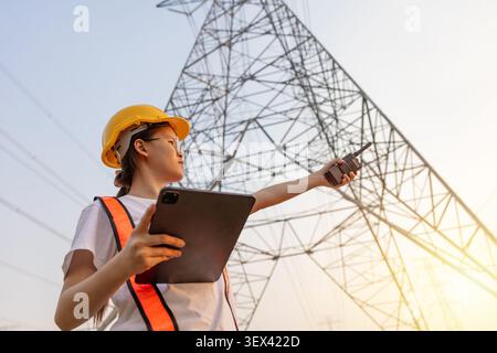 Female engineer inspecting power transmission tower, energy infrastructure concept, electricity industry, utility maintenance, engineering inspection, Stock Photo