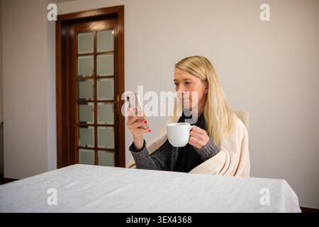 Woman relaxing at home, holding a warm drink and looking at mobile phone Stock Photo