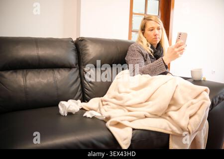 Sick woman feeling unwell, wrapped in blanket, using smartphone for online doctor consultation Stock Photo