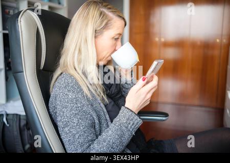Woman sitting in office chair, drinking from cup, and watching smartphone Stock Photo