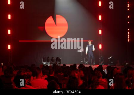 Business presenter on stage addresses seated audience under dramatic red lighting, logo projection visible on backdrop, conference atmosphere, public speaking, live event. Stock Photo