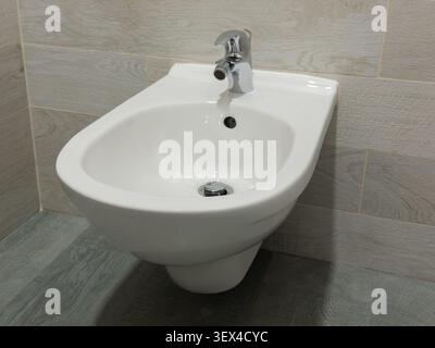 This minimalist white bidet installation mounted on a light tiled wall in a sleek bathroom emphasizes functional design and comfort for a sophisticate Stock Photo