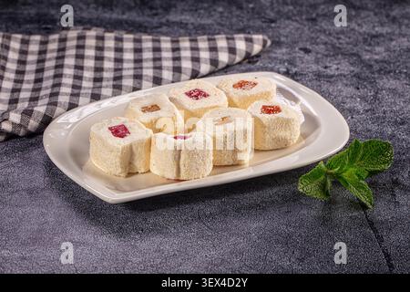 White coconut coated marshmallows with red orange fruit centers arranged on oval white plate. Fresh mint sprig beside plate. Dark textured surface bla Stock Photo