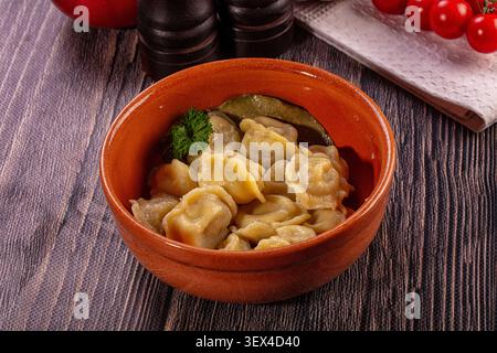 Traditional Russian dumplings pelmeni served in rustic clay bowl with parsley garnish and bay leaf, set on wooden table with tomatoes and pepper mills Stock Photo