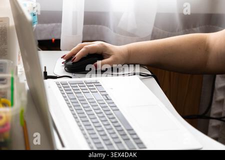 A close view of a hand with red nails operating a wired mouse next to a notebook in a light room, illustrating focused computer interaction for busine Stock Photo