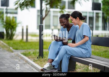 Two diverse healthcare workers enjoying a coffee break while looking at a smartphone Stock Photo