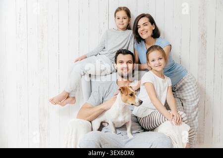 Family portrait. Happy parents with their two daughters and dog pose together against white background, spend free time at home, being in good mood. Mother, father and small sisters pose indoor Stock Photo