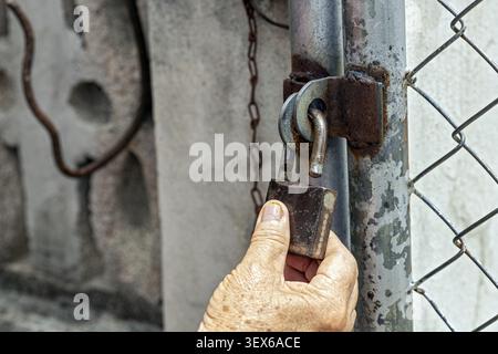 Close up of an elderly wrinkled hand holding an old rusty padlock attached to a corroded metal gate post, with chain link fence and weathered concrete Stock Photo