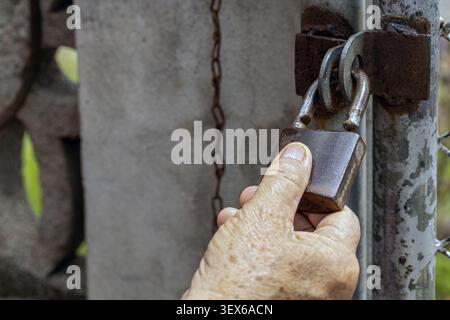 Close up of an elderly wrinkled hand holding a rusty brown padlock attached to a weathered metal gate latch with a hanging iron chain in the backgroun Stock Photo