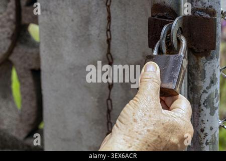 Wrinkled elderly hand grips an old rusty padlock securing a weathered metal gate with hanging chain, conveying themes of security, property protection Stock Photo