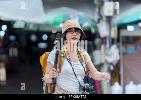 Asian woman tourist with backpack holding smartphone on selfie stick for travel vlog Stock Photo