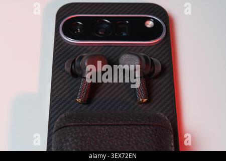 Close up high angle view of modern black wireless earbuds lying on the back of a smartphone in carbon fiber texture protective case. Stock Photo