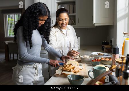 African American mom and teenage daughter making breakfast in kitchen slicing loaf on cutting board Stock Photo
