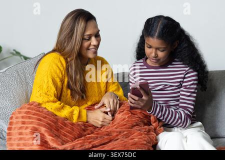 African mother and teenage daughter sitting on gray couch, holding smartphone, sharing blanket Stock Photo