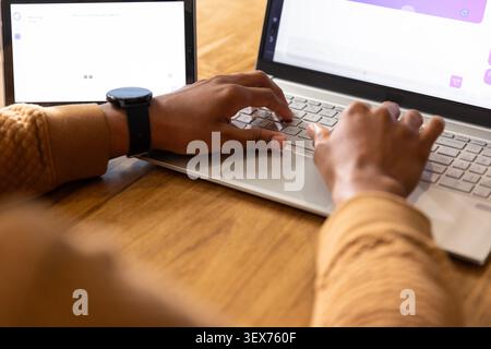 Man typing on silver laptop at wooden table, in tan knit top, black smartwatch, referencing tablet Stock Photo