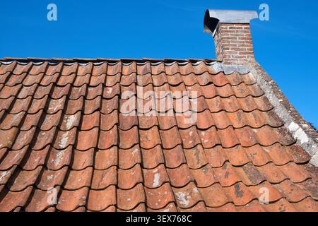 Weathered terracotta roof tiles with a brick chimney with a metal cap under a clear blue sky, of a 17th-century Dutch farmhouse Stock Photo