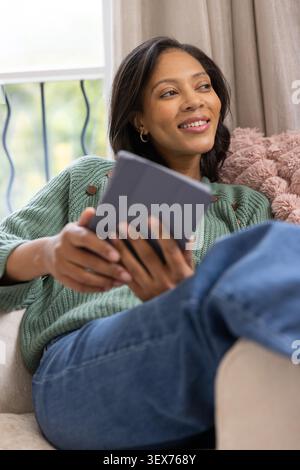 African American woman reclining on sofa near window, holding gray tablet and wearing green sweater Stock Photo