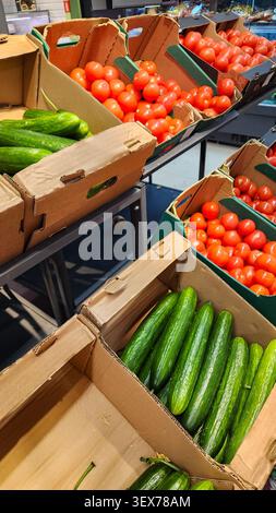 Assorted fresh red tomatoes, green cucumbers, displayed in cardboard boxes and plastic crates on a shelf inside a grocery store. Vertical photo Stock Photo
