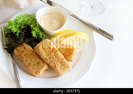 Crispy breaded fish fingers served with fresh green salad, lemon wedges, and mayonnaise dip on a white plate, white background. Copy space. Stock Photo