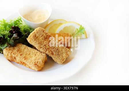 Crispy breaded fish fingers served with fresh green salad, lemon wedges, and mayonnaise dip on a white plate, white background. Copy space. Stock Photo