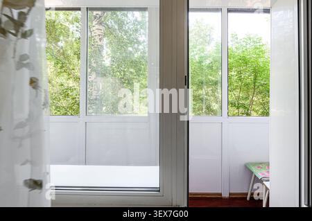 View of a balcony through an open doorway, with green trees visible through the windows. White frames and curtain enhance the bright, airy feel Stock Photo