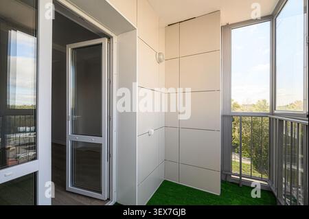 bright balcony space featuring white tiled walls, artificial grass, and an open glass door leading inside. Outside, greenery and trees are visible thr Stock Photo