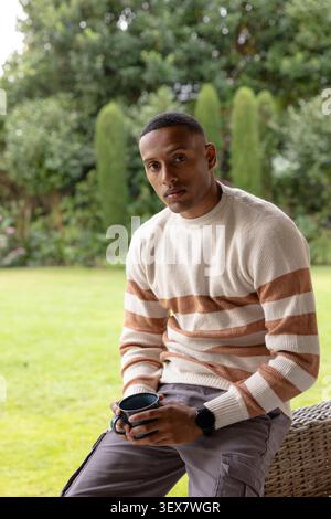 African man sitting on stool in backyard, wearing cream sweater and grey cargo pants, holding mug Stock Photo