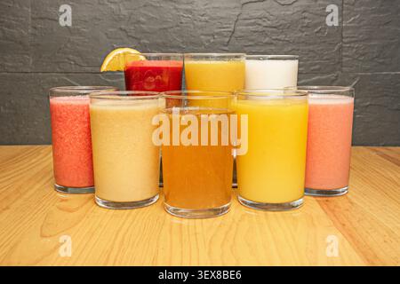Assortment of colorful fresh fruit juices and smoothies in clear glasses arranged on a wooden table against a dark textured stone wall background Stock Photo