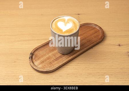 Hot cappuccino featuring a perfect heart-shaped latte art design served in a rustic grey ceramic cup placed on an oval wooden tray atop a light table Stock Photo
