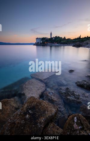 Long exposure landscape of historical monastery on Adriatic sea coast. Beautiful twilight sky over calm water bay with rocks in Bol, Croatia. Stock Photo