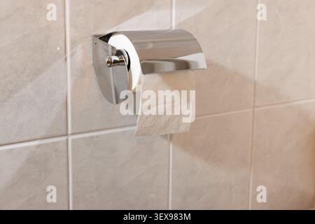 Modern chrome toilet roll holder mounted on beige tiled wall. Paper partially unrolled, neatly placed. Simple bathroom design, minimalist aesthetic. I Stock Photo
