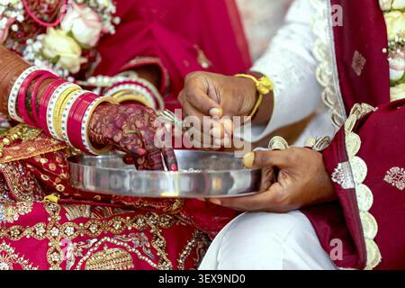 Close-up of a bride's intricately henna-decorated hand adorned with vibrant bangles, and a groom's hand holding rice grains during a traditional India Stock Photo