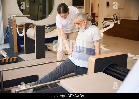 Elderly man stretching leg with assistance from young woman in clinic Stock Photo