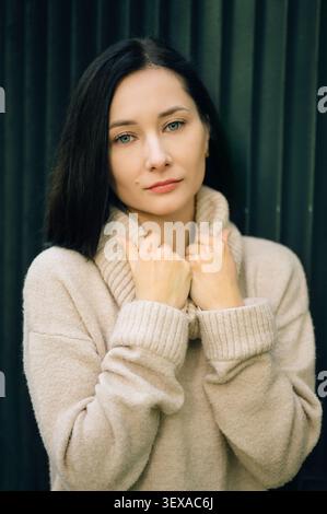 Beautiful brunette woman holding the collar of her beige sweater against a dark ribbed background Stock Photo